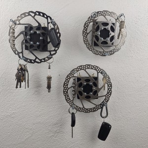 Könnte beinhalten: Drei metallene Schlüsselhalter in Form von Scheiben, die an einer weißen Wand montiert sind. Jede Scheibe hat ein schwarzes Quadrat in der Mitte mit einem Lochmuster. An den Scheiben hängen verschiedene Schlüssel, eine Zündkerze und ein Autoschlüssel.