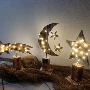 Könnte beinhalten: Drei hölzerne Sternen- und Halbmond-Nachtlichter mit warmweißen LED-Lichtern. Die Lichter stehen auf Holzsockeln und sind auf einer braunen Stoffoberfläche angeordnet.