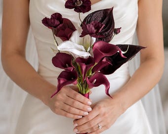Puede incluir: Un ramo de flores con calas de color burdeos intenso, amapolas de color púrpura oscuro y una sola cala blanca. Los tallos están unidos y envueltos con una cinta de color burdeos a juego, creando un contraste con la tela blanca de fondo.