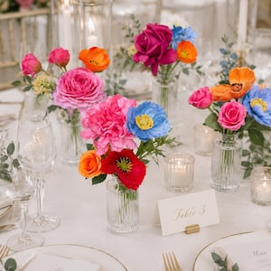 May include: A table setting featuring multiple small, clear, ribbed glass vases filled with vibrant artificial flowers in shades of hot pink, bright orange, red, and blue. A white place card with the text Table 3 sits near gold-toned cutlery.