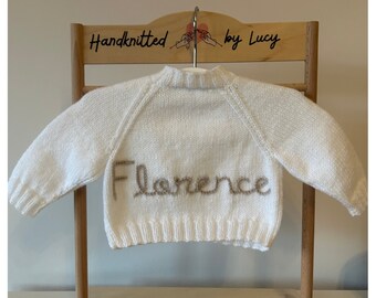 May include: White hand-knitted baby sweater with long sleeves and a crew neck. The sweater has the name "Florence" embroidered on the front in a light brown yarn.