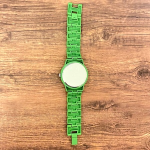 May include: A bright green wristwatch with a matching band. The watch face is round with a reflective surface. The band is made of rectangular links. The watch is set against a wooden background.