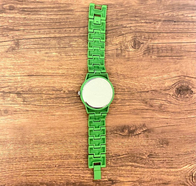 May include: A bright green wristwatch with a matching band. The watch face is round with a reflective surface. The band is made of rectangular links. The watch is set against a wooden background.