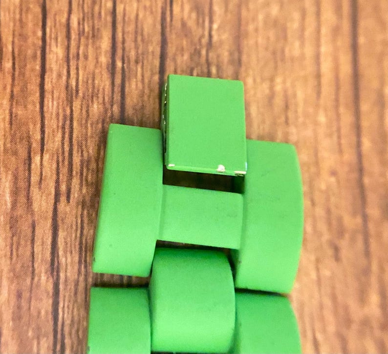 May include: Close-up of a bright green watch band, featuring rectangular and curved links. The band is made of a solid, vibrant green material, with a few minor imperfections. The background is a wooden surface.