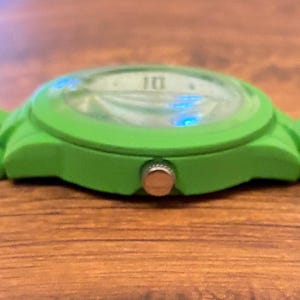 May include: A close-up of a bright green wristwatch. The watch face is visible, displaying the number 10. The watch has a green band and a silver-colored button on the side. The watch is resting on a wooden surface.