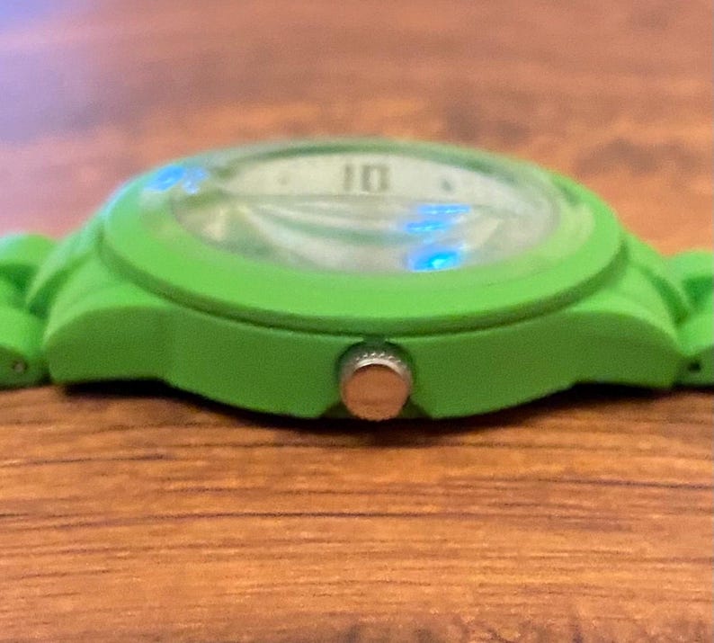 May include: A close-up of a bright green wristwatch. The watch face is visible, displaying the number 10. The watch has a green band and a silver-colored button on the side. The watch is resting on a wooden surface.