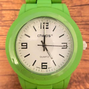 May include: A bright green wristwatch with a white face. The watch face features black numerals at the 12, 3, 6, and 9 o'clock positions, with black dashes marking the other hours. The brand name "chico's&reg;" is printed on the face.
