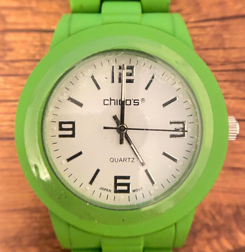 May include: A bright green wristwatch with a white face. The watch face features black numerals at the 12, 3, 6, and 9 o'clock positions, with black dashes marking the other hours. The brand name "chico's&reg;" is printed on the face.