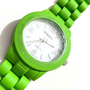 May include: Bright green quartz watch with a matching green band. The watch face is white with silver hands and the brand name "CHICO'S". The watch has a round face and a textured band.