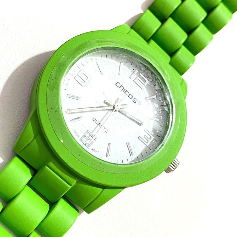 May include: Bright green quartz watch with a matching green band. The watch face is white with silver hands and the brand name "CHICO'S". The watch has a round face and a textured band.