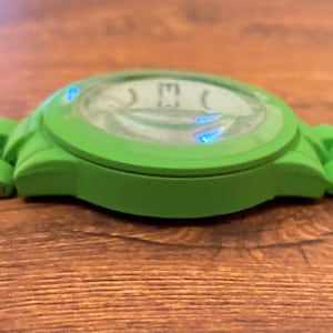 May include: A green wristwatch with a round face and a matching green band. The watch face has a light-colored background with black numerals. The watch is made of a flexible material and is set against a wooden surface.