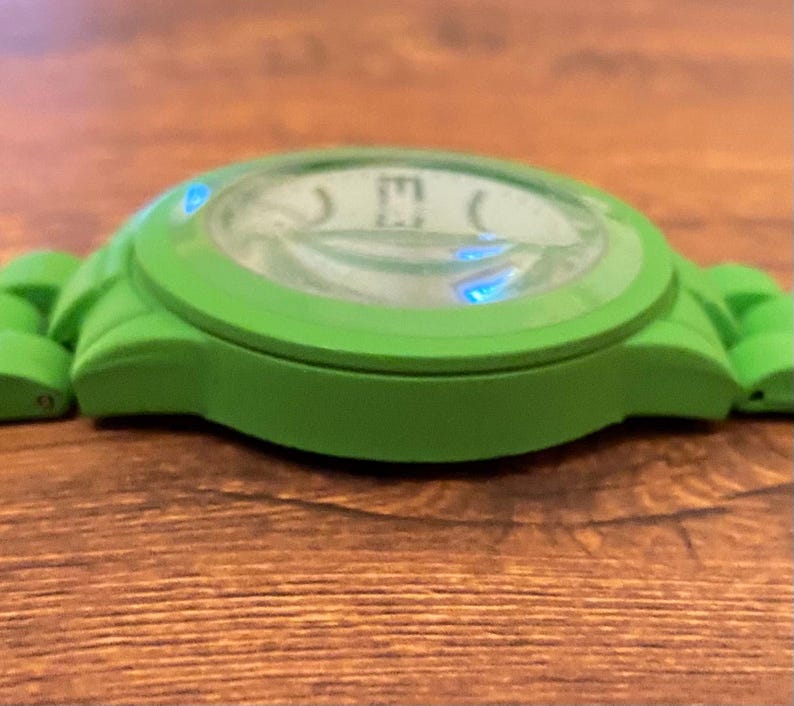 May include: A green wristwatch with a round face and a matching green band. The watch face has a light-colored background with black numerals. The watch is made of a flexible material and is set against a wooden surface.
