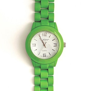 May include: A bright green wristwatch with a white watch face. The watch features a round face with black numerals and hands, and the brand name "NICO'S" is printed on the face. The watch band is also bright green.
