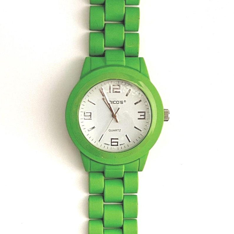 May include: A bright green wristwatch with a white watch face. The watch features a round face with black numerals and hands, and the brand name "NICO'S" is printed on the face. The watch band is also bright green.