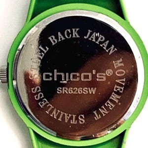 May include: Close-up of the back of a green watch with a green band. The watch face is silver with the words "STEEL BACK JAPAN MOVEMENT STAINLESS" around the edge. The brand name "chico's" is in the center.