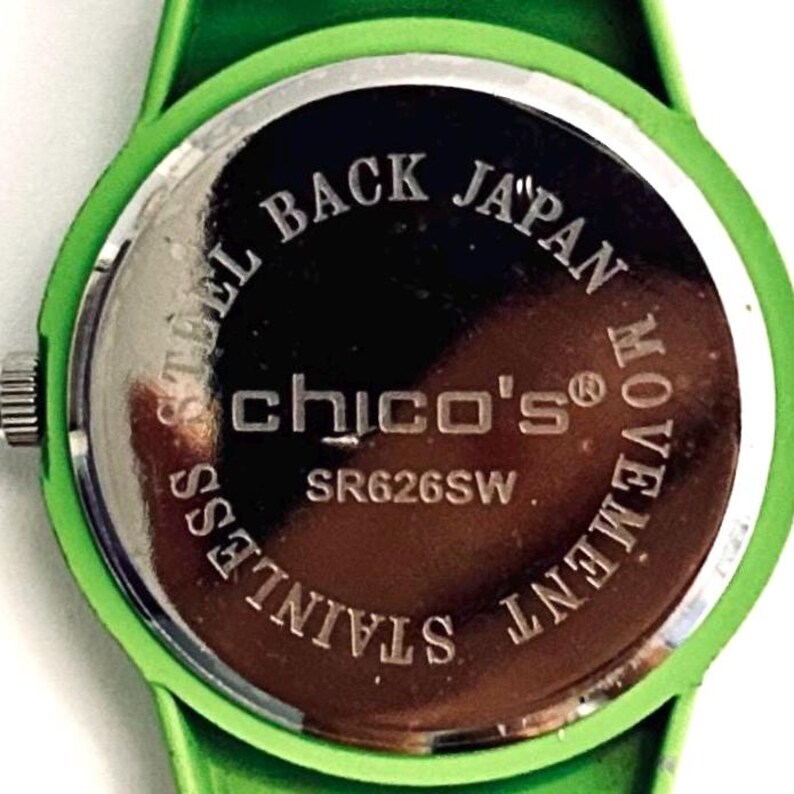 May include: Close-up of the back of a green watch with a green band. The watch face is silver with the words "STEEL BACK JAPAN MOVEMENT STAINLESS" around the edge. The brand name "chico's" is in the center.