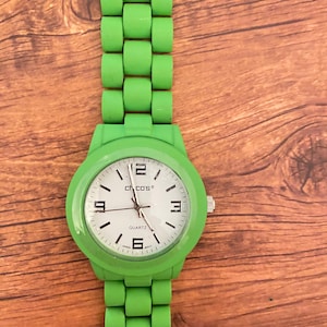 May include: A bright green wristwatch with a matching band. The watch face is white with black numerals and hands, and the brand name "Chicos" is visible. The watch band is made of a series of connected, rounded blocks.