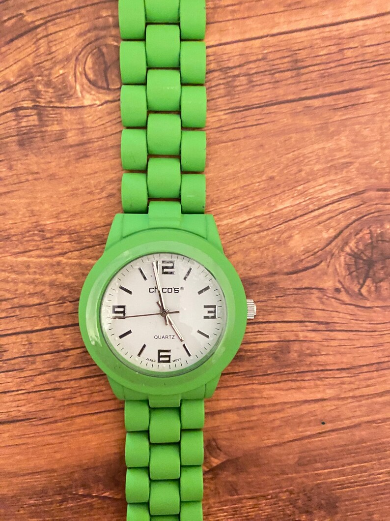 May include: A bright green wristwatch with a matching band. The watch face is white with black numerals and hands, and the brand name "Chicos" is visible. The watch band is made of a series of connected, rounded blocks.