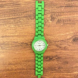 May include: A bright green wristwatch with a matching band. The watch face is white with black numerals and hands. The brand name "Chicos" is printed on the watch face. The watch is displayed on a wooden surface.