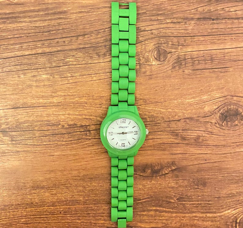 May include: A bright green wristwatch with a matching band. The watch face is white with black numerals and hands. The brand name "Chicos" is printed on the watch face. The watch is displayed on a wooden surface.