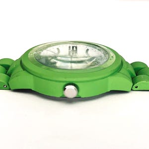 May include: A bright green wristwatch with a matching band. The watch face is clear with silver-colored hands and markers. The watch case and band are a solid, vibrant green color, creating a bold and modern look. The watch is designed with a simple, minimalist aesthetic.