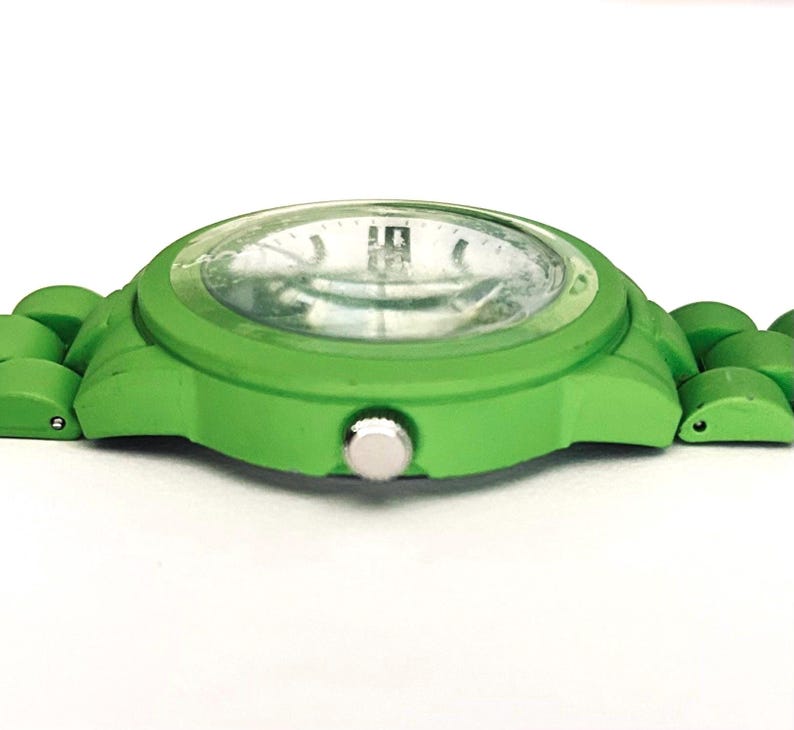 May include: A bright green wristwatch with a matching band. The watch face is clear with silver-colored hands and markers. The watch case and band are a solid, vibrant green color, creating a bold and modern look. The watch is designed with a simple, minimalist aesthetic.