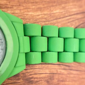 May include: Close-up of a bright green watch with a matching band. The band features a unique cylindrical design. The watch face is partially visible, showing a white dial with black markings. The watch is set against a wooden background.