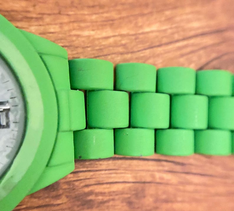 May include: Close-up of a bright green watch with a matching band. The band features a unique cylindrical design. The watch face is partially visible, showing a white dial with black markings. The watch is set against a wooden background.