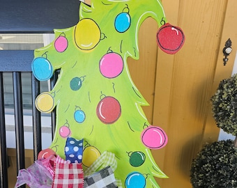 May include: A green wooden Christmas tree cutout with a brown base. The tree is decorated with colorful ornaments and a multi-colored bow.