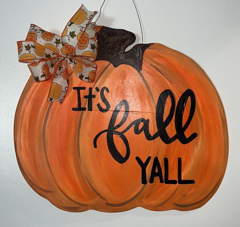 May include: A wooden pumpkin door hanger painted orange with brown details. It has a brown stem and a white and orange ribbon bow. The text "It's fall y'all" is painted in black on the pumpkin.
