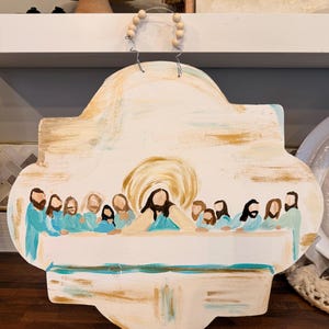 May include: Hand-painted wooden wall hanging depicting the Last Supper. The artwork features figures in blue and beige robes around a table, with a golden halo around the central figure. The sign has a decorative shape and a wire hanger with wooden beads.
