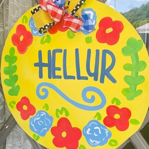 May include: A yellow round wooden sign with a blue floral design and the word "HELLUR" written in blue paint. The sign has a red and white gingham bow with black polka dots.