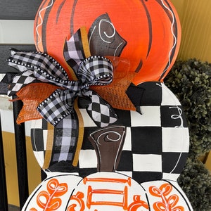 May include: A three-tiered pumpkin door hanger with a black and white checkered pattern, a black and white ribbon bow, and the words "Give Thanks" in orange on the bottom pumpkin.