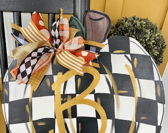 May include: A black and white chequered pumpkin door hanger with a gold letter "B" in the centre. The pumpkin has a brown stem and is decorated with a gold and black bow.