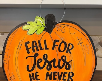 May include: Orange pumpkin-shaped door hanger with a black stem and green leaf. The pumpkin has black lettering that reads "Fall for Jesus He Never Leaves".