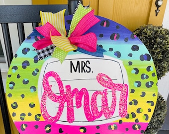 May include: A round wooden door hanger with a rainbow of colours and a leopard print pattern. The door hanger has a white circle with the text "MRS. Smart" in pink glitter letters.