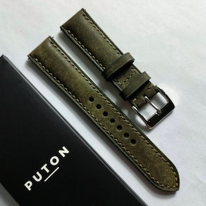 Può includere: Cinturino per orologio in pelle verde oliva con fibbia argentata. Il cinturino ha una finitura invecchiata e cuciture a vista. Una scatola nera con la scritta "PUTON" in lettere bianche è parzialmente visibile.