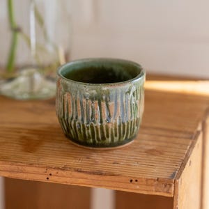Op de afbeelding: Een klein, groen keramiek kopje met een textuur, druipende glazuur. Het kopje staat op een houten oppervlak.