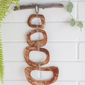 May include: A wall hanging made of four brown ceramic rings, each with a different size and shape, connected by twine. The rings are hanging from a piece of driftwood with gold beads.