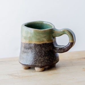 May include: A small, handmade ceramic mug with a green, brown and black glaze. The mug has a rounded body and a handle. It sits on four small feet.