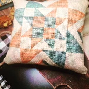 May include: A handmade pillow with a geometric pattern in shades of peach, teal, and white. The pillow is made of a soft, textured fabric and has a square shape.