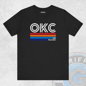 Op de afbeelding: Zwart T-shirt met de witte tekst "OKC" boven horizontale strepen in oranje, rood, blauw en marine. Het woord "thunder" staat eronder. Het shirt is van het merk Black-Tee.