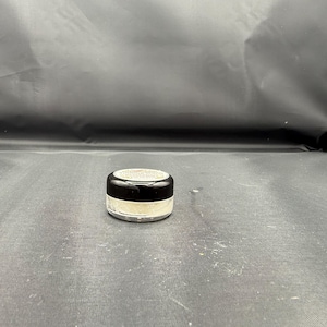 Peut inclure: Un petit pot cosmétique rond en verre transparent avec un couvercle noir à vis. Le récipient contient une substance poudreuse de couleur claire visible à travers la base transparente. Le couvercle porte une étiquette circulaire avec du texte.