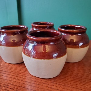 Könnte beinhalten: Vier braune und weiße Keramikkrüge mit glasierter Oberfläche. Die Krüge sind in einer Reihe auf einer Holzoberfläche angeordnet.