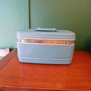 May include: A vintage blue vanity case with a silver-toned accent band. The rectangular case has a handle on top and is set on a wooden surface. The case has a textured surface and silver trim.