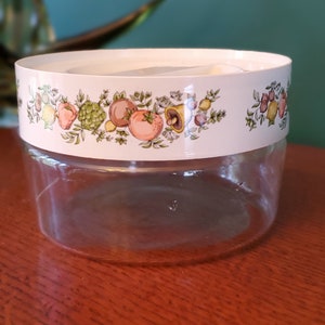 Puede incluir: Un tarro de vidrio transparente con una tapa blanca y un diseño floral colorido con frutas y verduras. La tapa tiene un patrón floral verde, amarillo, naranja y rosa.
