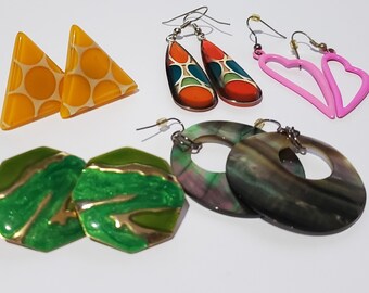 Op de afbeelding: Assortiment van kleurrijke oorbellen, waaronder een paar gele driehoekige oorbellen met witte stippen, een paar groene zeshoekige oorbellen met gouden accenten, een paar ronde oorbellen met een zwart-wit marmerpatroon, een paar hartvormige oorbellen in roze en een paar druppelvormige oorbellen met een veelkleurig geometrisch patroon.