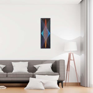 May include: A rectangular wall art piece with a geometric design. The artwork features a diamond shape with blue, orange, and yellow threads against a black background. The piece is mounted on a white wall above a gray sofa.