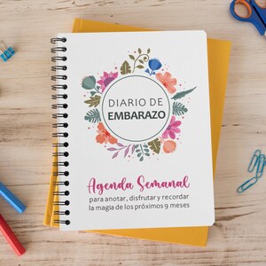 May include: A white spiral bound journal with a floral design and the text "DIARIO DE EMBARAZO" in a circle. The text "Agenda Semanal" is below the circle with the text "para anotar, disfrutar y recordar la magia de los próximos 9 meses".