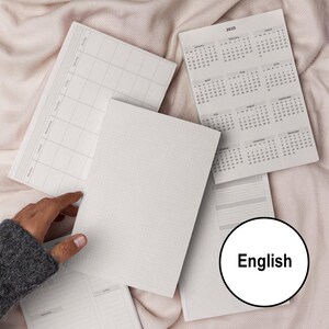 Puede incluir: Un conjunto de cuadernos en blanco con páginas cuadriculadas y rayadas. Los cuadernos son blancos con una textura sutil. La imagen también incluye un calendario para el año 2025. El calendario está impreso en una hoja de papel blanca con texto negro. El texto "English" está impreso en negro sobre un círculo blanco.